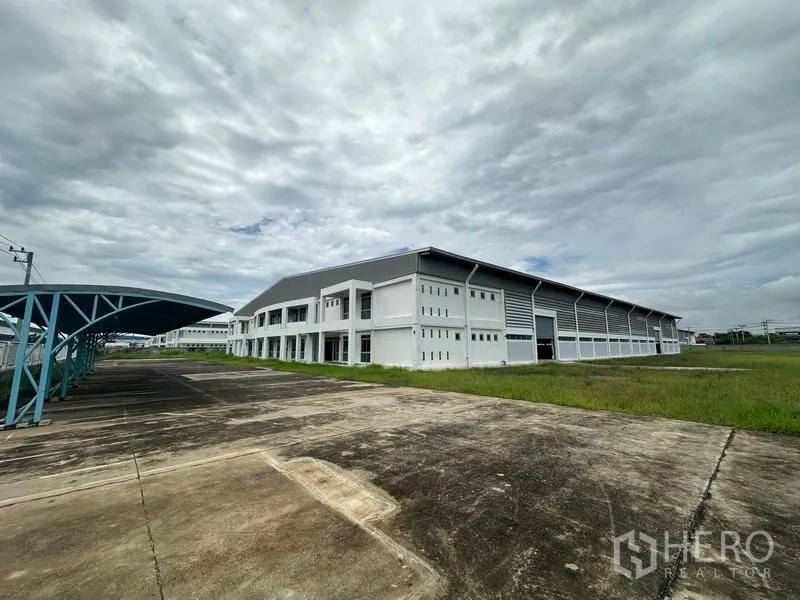 工厂 出租在Pathum Thani - 现代厂房外观，宽阔车道与带顶棚停车位，阴天。