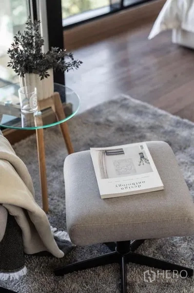 Condominium for rent or sale in Phra Khanong, Bangkok - Reading nook with ottoman, glass side table and magazine on a gray rug near a window.