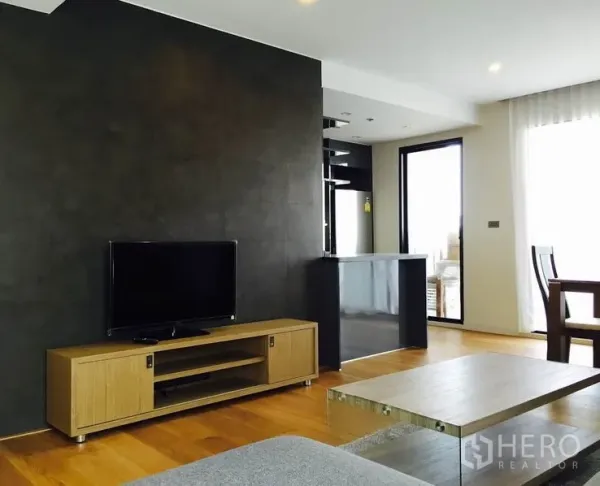 คอนโด ให้เช่าในWatthana, Bangkok - ห้องนั่งเล่นพื้นไม้ พร้อมชั้นวางทีวีและผนังสีเข้ม เชื่อมต่อครัวและประตูกระจกออกสู่ระเบียง