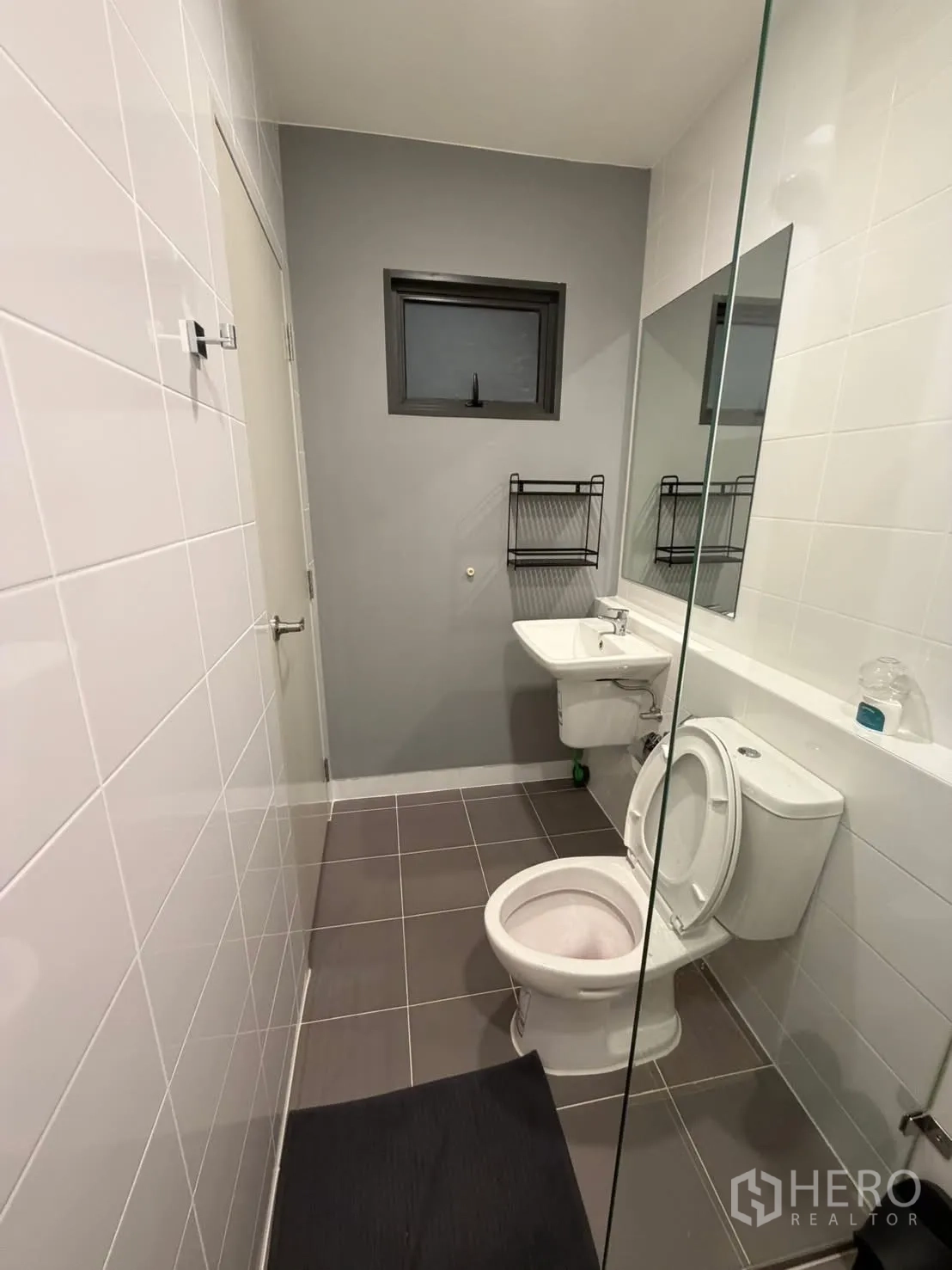 Townhouse for rent in Bang Phli, Samut Prakan - Bathroom with grey and white tiles, wall-mounted sink, toilet, small window and glass divider.