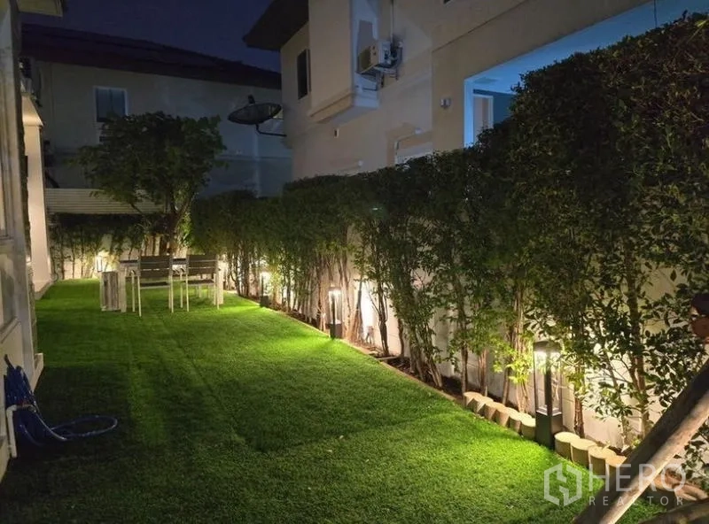 独立式住宅 出租 - 侧院夜景，整齐绿篱、庭院灯和户外餐桌。