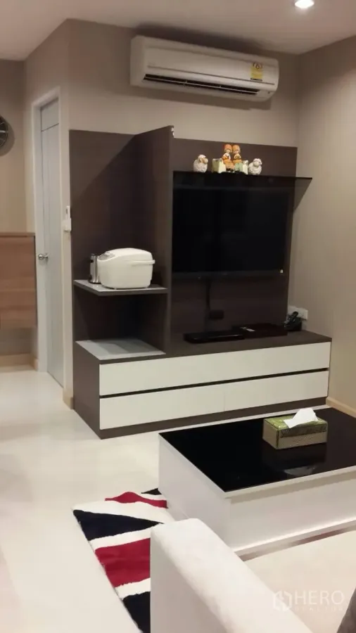 Condominium for rent in Mueang Samut Prakan, Samut Prakan - Contemporary living area with wall-mounted TV, built-in storage, air conditioning, and a glossy coffee table over a patterned rug.