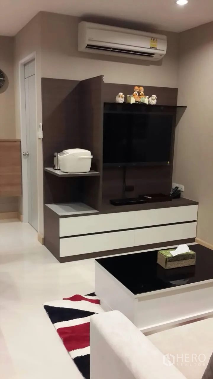 Condominium for rent in Mueang Samut Prakan, Samut Prakan - Contemporary living area with wall-mounted TV, built-in storage, air conditioning, and a glossy coffee table over a patterned rug.