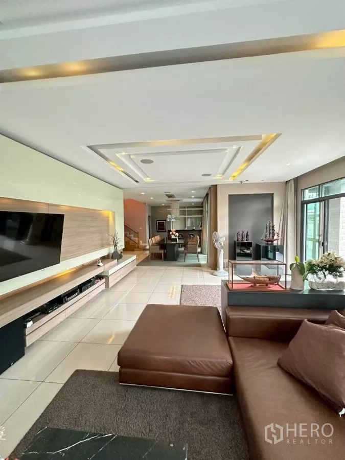 Detached House for sale in Bang Lamung, Chon Buri - Spacious living room with brown sectional sofa, TV wall and recessed ceiling lights.