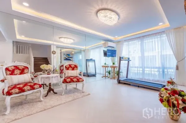 联排别墅 出租在Lat Phrao, Bangkok - 明亮客厅，整面镜墙与水晶灯，红色古典单椅，落地窗旁有跑步机。