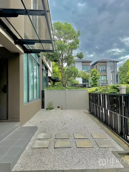 บ้าน ให้เช่าหรือขายในPrawet, Bangkok - ลานหน้าบ้านโรยกรวด หลังคากันสาดกระจก และรั้วเหล็กหันสู่ถนนในโครงการที่ร่มรื่น