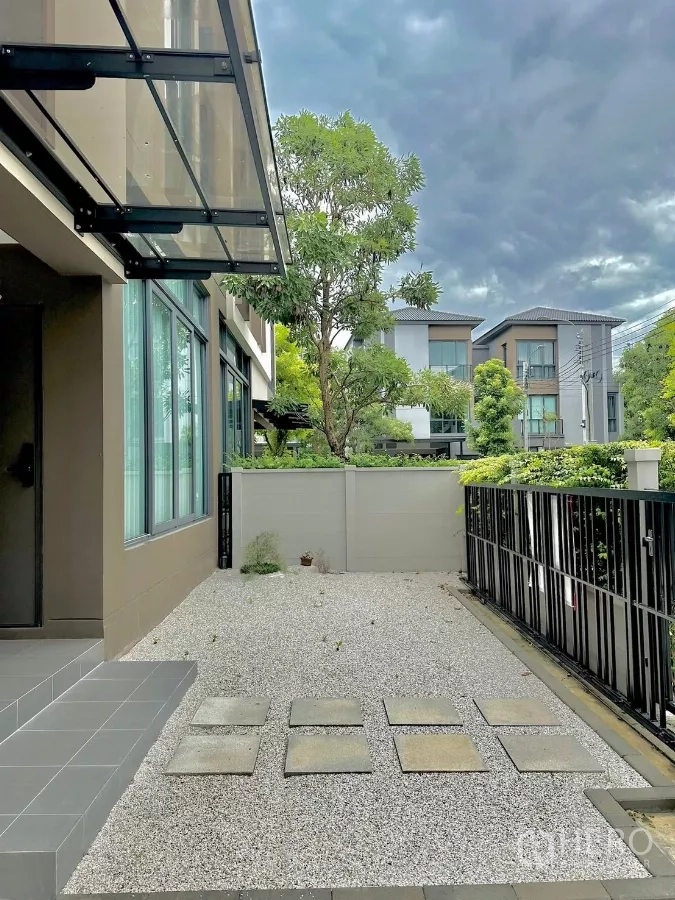 บ้าน ให้เช่าหรือขายในPrawet, Bangkok - ลานหน้าบ้านโรยกรวด หลังคากันสาดกระจก และรั้วเหล็กหันสู่ถนนในโครงการที่ร่มรื่น