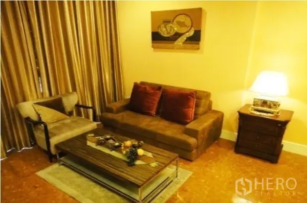 Condominium for rent in Khlong Toei, Bangkok - Warm-toned living room with sofa, armchair, coffee table, wall art, and floor-to-ceiling curtains.