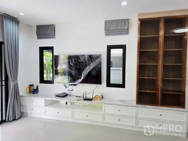 Detached House for rent in Prawet, Bangkok - Living room feature wall with built-in TV cabinet, white drawers, glass display unit and grey curtains.