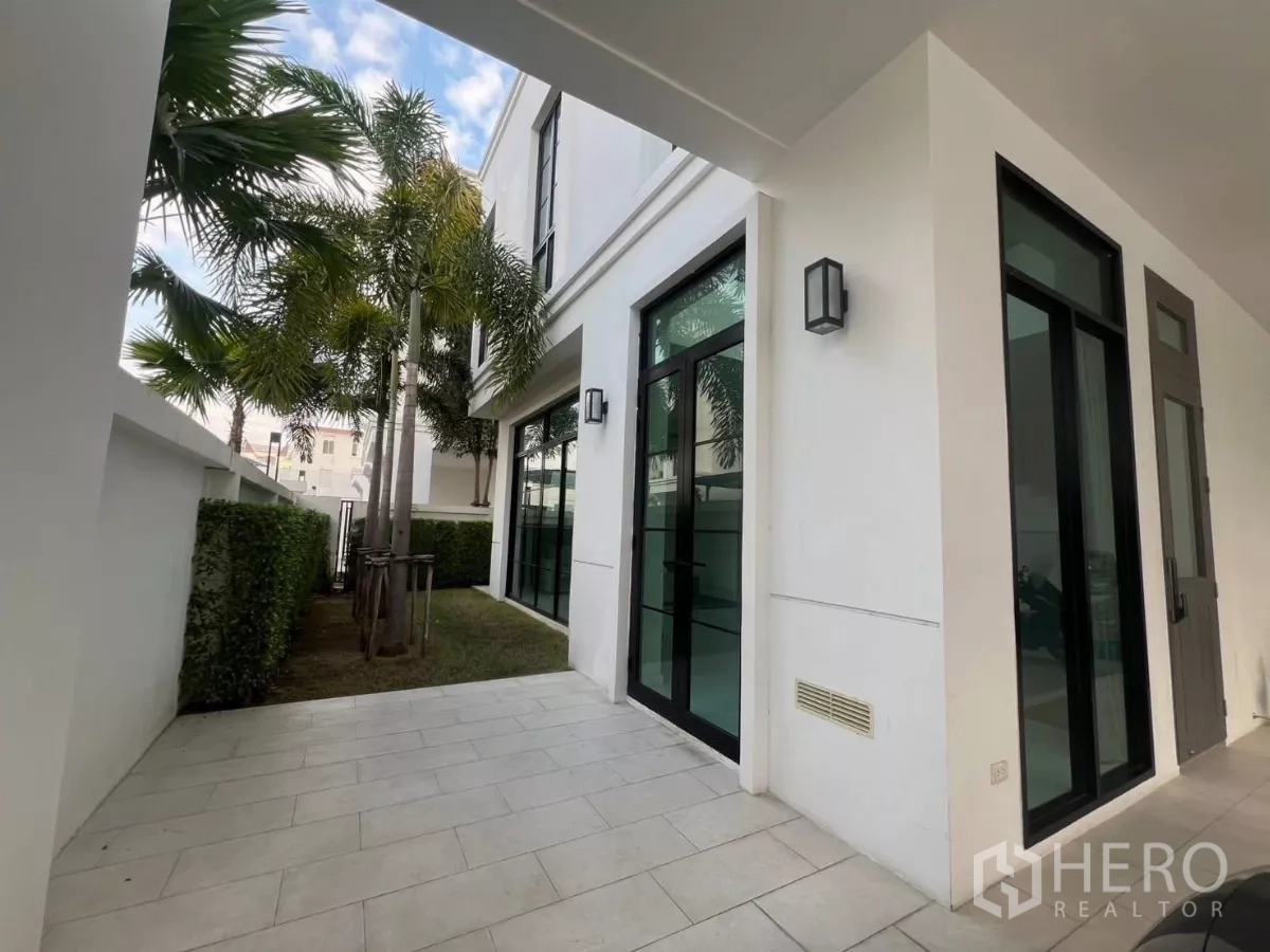 บ้าน ให้เช่าในLat Phrao, Bangkok - เฉลียงข้างบ้านและสนามหญ้าขนาดกะทัดรัด มีต้นปาล์ม แนวรั้วพุ่มเตี้ย และประตูกระจกกรอบอลูมิเนียมสีดำ