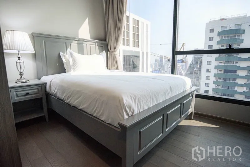 Condominium for rent or sale in Watthana, Bangkok - Secondary bedroom with gray panel bed, corner window and urban view.