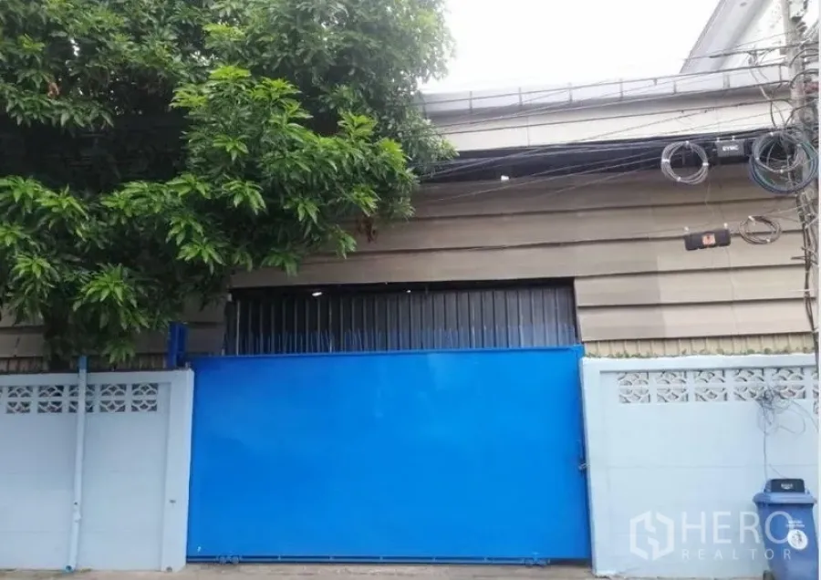 Warehouse for rent in Bang Kapi, Bangkok - Secure blue gate and low-rise warehouse facade shaded by a large tree.