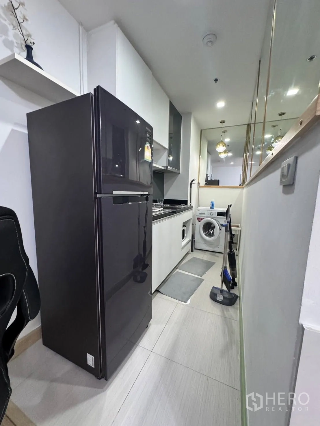 Condominium for rent in Watthana, Bangkok - Compact galley kitchen with black refrigerator, cabinets, sink, and washing machine.