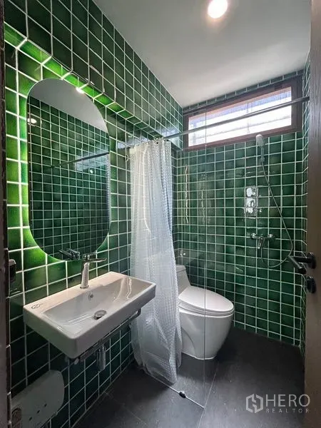 Apartment for rent in Khlong Toei, Bangkok - Compact bathroom with green tiled walls, wall-mounted sink, toilet and glass-partition shower with a window.