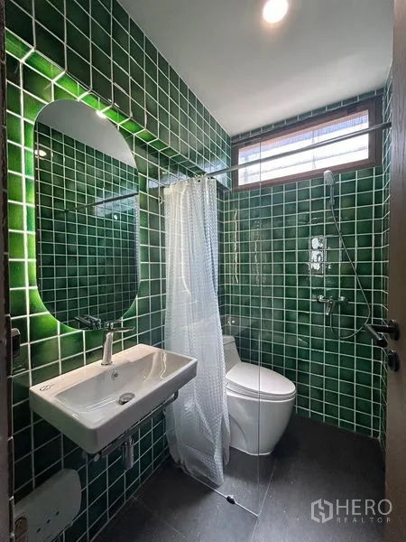 Apartment for rent in Khlong Toei, Bangkok - Compact bathroom with green tiled walls, wall-mounted sink, toilet and glass-partition shower with a window.