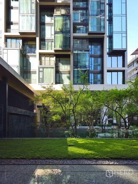 Condominium for rent or sale in Pathum Wan, Bangkok - Modern condominium facade with expansive glass windows overlooking a landscaped lawn and trees.