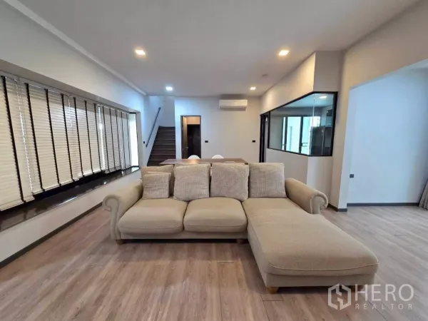 Detached House for rent in Bang Phli, Samut Prakan - Bright living room with L-shaped sofa, wood floors, staircase and a glass-partitioned kitchen.