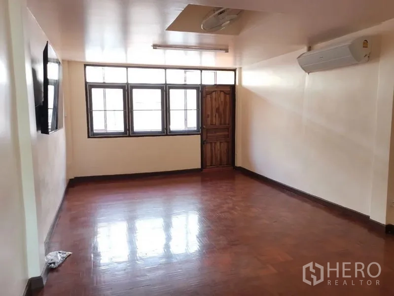 Townhouse for rent in Watthana, Bangkok - Spacious living area with polished wooden floor, multiple windows, and wall-mounted air conditioner.