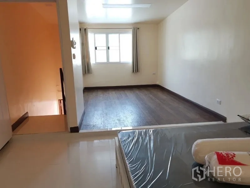 Townhouse for rent in Watthana, Bangkok - Large bedroom with step-up wood flooring, window with curtains, and new mattress wrapped in plastic.