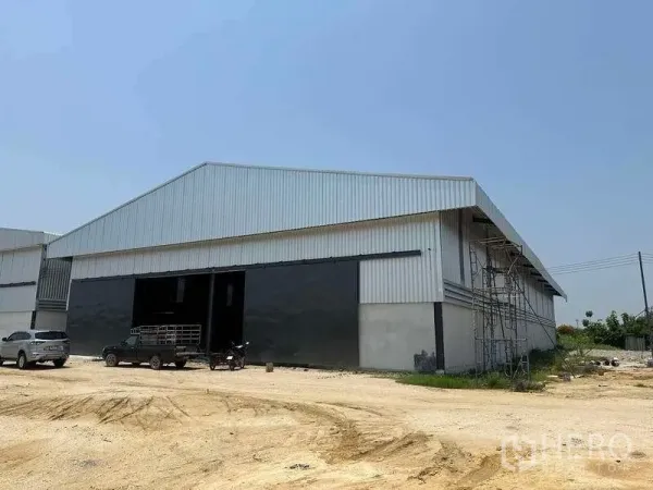 工厂 出租在Phan Thong, Chon Buri - 工厂斜角外观，金属板墙体、超大门、脚手架与开阔空地。