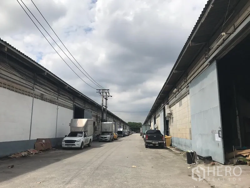 คลังสินค้า ให้เช่าในBang Phli, Samut Prakan - โกดังเรียงรายสองฝั่งถนนภายในกว้าง รถบรรทุกจอดอยู่ใต้ท้องฟ้ามีเมฆ