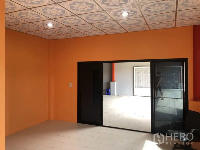 货栈 出租在Nonthaburi - 底层办公室，橙色墙面、花纹吊顶，黑色推拉玻璃门通往仓库。