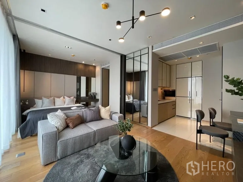 Condominium for sale - Open-plan living room with grey sofa, glass coffee table, compact kitchen and a partitioned bedroom.