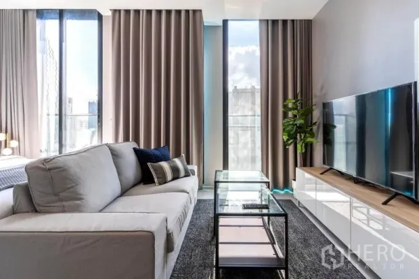 คอนโด ให้เช่าในPathum Wan, Bangkok - มุมนั่งเล่นสว่างด้วยโซฟาสีเทา โต๊ะกลางกระจก ตู้วางทีวี และหน้าต่างสูงจรดเพดานมองวิวเมือง