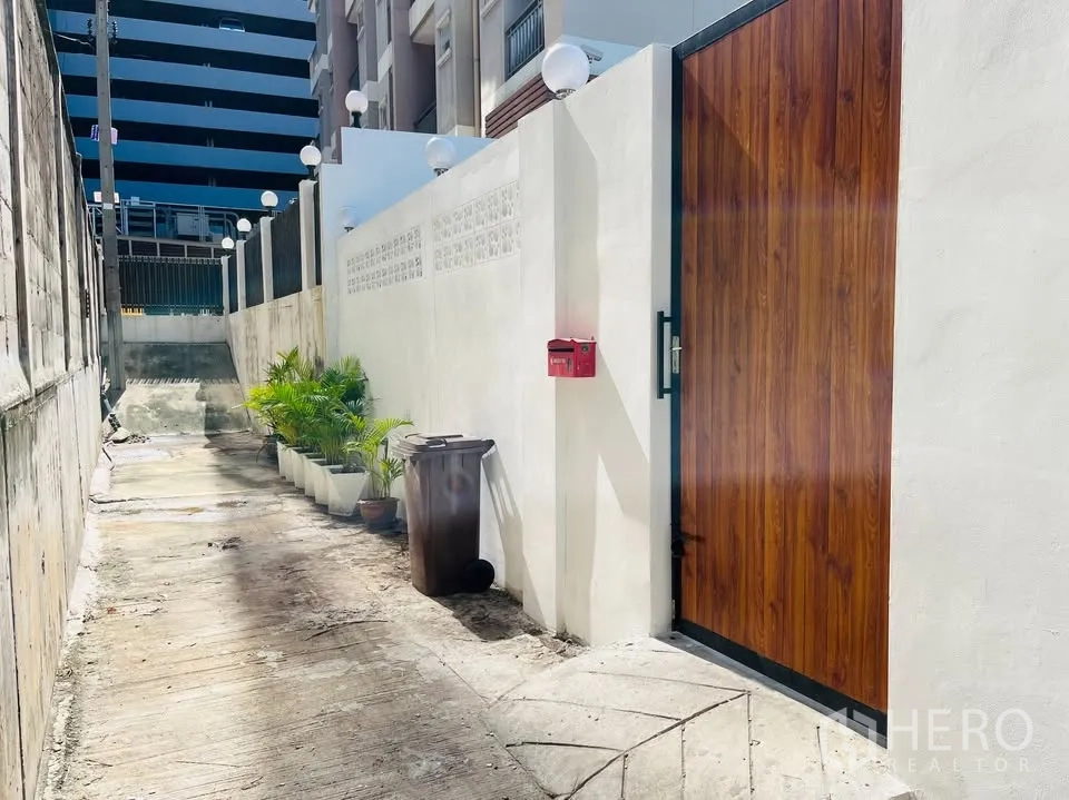 บ้าน ให้เช่า - ทางเข้าซอยแคบมีกำแพงสีขาวและประตูไม้สูง พร้อมต้นไม้กระถาง