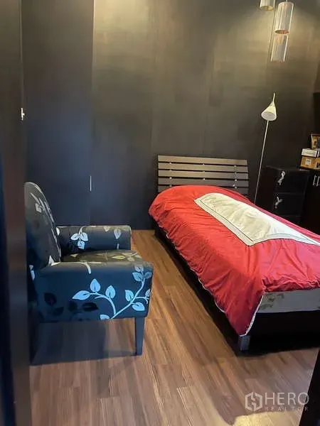 Detached House for sale in Suan Luang, Bangkok - Compact bedroom with a single bed in a red duvet, armchair and wood flooring against dark walls.