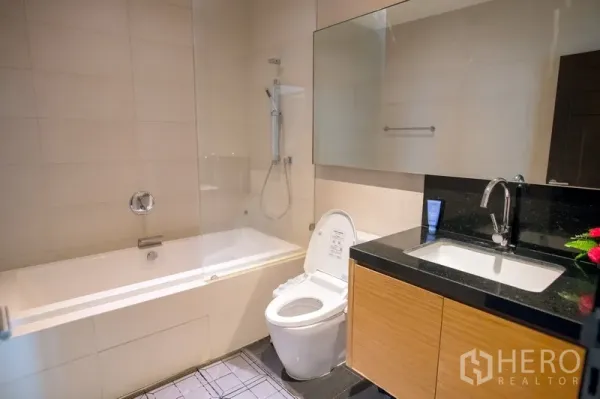 Condominium for rent in Watthana, Bangkok - Modern bathroom featuring a bathtub with shower, glass screen, large mirror, and vanity with black countertop.