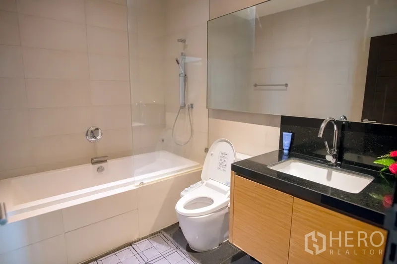 Condominium for rent in Watthana, Bangkok - Modern bathroom featuring a bathtub with shower, glass screen, large mirror, and vanity with black countertop.