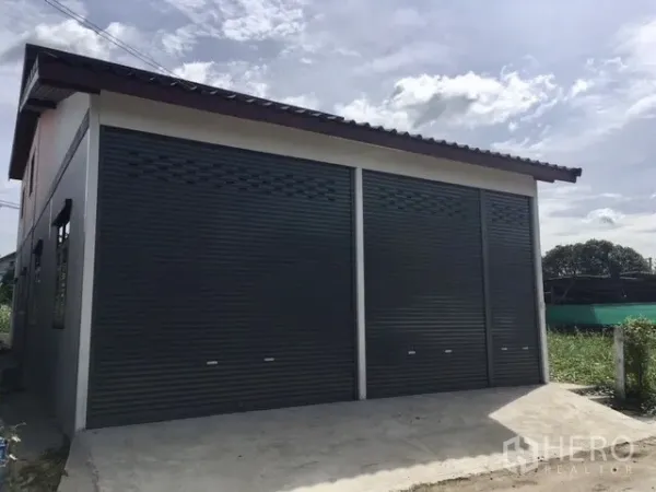 คลังสินค้า ให้เช่าในLam Luk Ka, Pathum Thani - ด้านหน้าโกดังสมัยใหม่ ประตูม้วนเหล็ก 2 ช่อง พร้อมทางเข้าคอนกรีตและหลังคาทรงจั่ว.