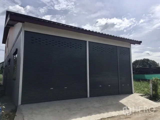 คลังสินค้า ให้เช่าในLam Luk Ka, Pathum Thani - ด้านหน้าโกดังสมัยใหม่ ประตูม้วนเหล็ก 2 ช่อง พร้อมทางเข้าคอนกรีตและหลังคาทรงจั่ว.