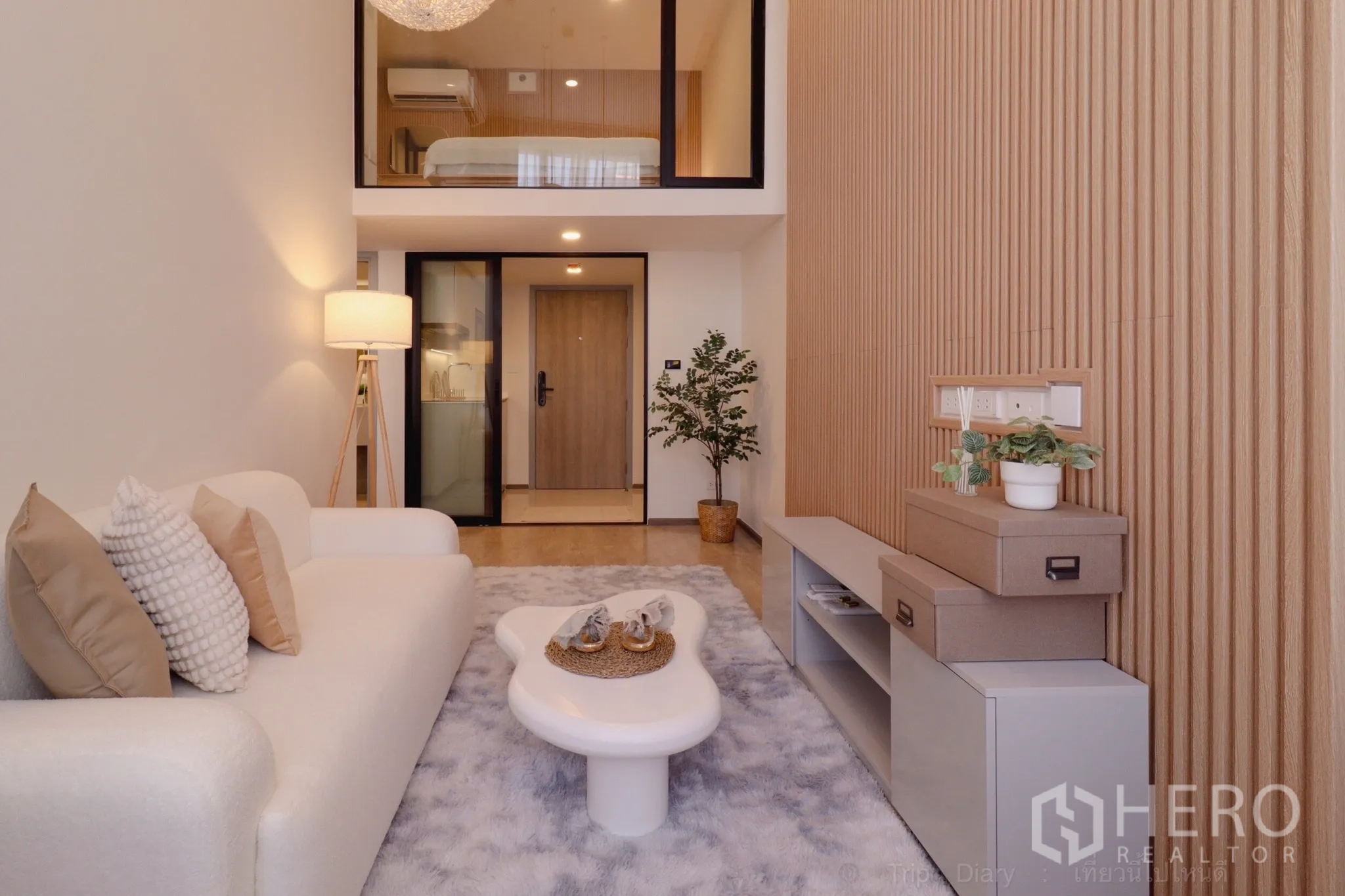 Condominium for rent in Huai Khwang, Bangkok - Muji-style loft living room with 4m ceiling, white sofa, sculptural coffee table, wood slat wall, mezzanine bedroom, and kitchenette behind glass.