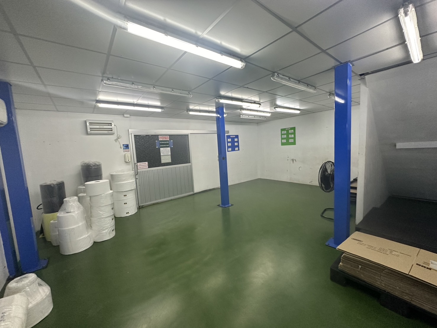 货栈 出租或出售在Lam Luk Ka, Pathum Thani - 开放式作业区，蓝色钢柱、空调与包装材料卷放在绿色地面上。