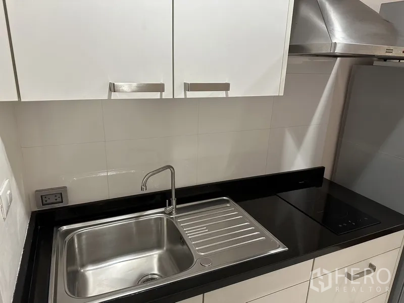 Condominium for rent in Watthana, Bangkok - Compact kitchen with stainless sink, black stone countertop and white cabinets.