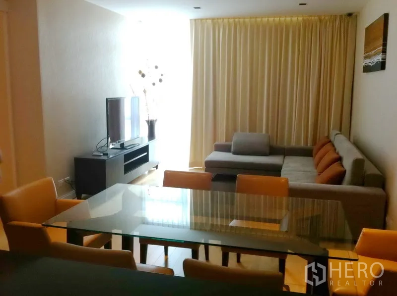 Condominium for rent in Pathum Wan, Bangkok - Open-plan living and dining area with glass table, L-shaped sofa, TV console, and floor-to-ceiling curtains.
