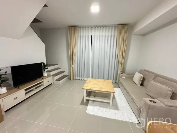 ทาวน์เฮาส์ ให้เช่าในBang Phli, Samut Prakan - ห้องนั่งเล่นสว่าง โซฟาสีเบจ ชั้นวางทีวี บันได และผ้าม่านสูงจรดเพดาน