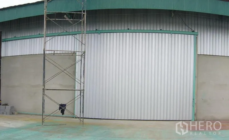 货栈 出租在Mueang Pathum Thani, Pathum Thani - 仓库外立面，宽大的金属推拉门与青绿色屋檐，前方为混凝土地面。