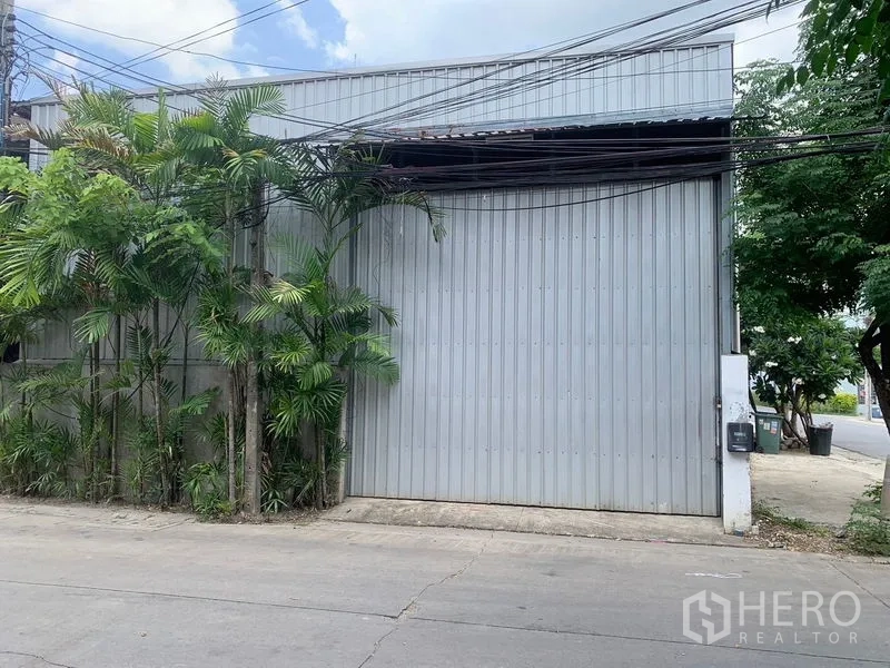 คลังสินค้า ให้เช่าในBang Kapi, Bangkok - ภาพระยะใกล้ประตูเมทัลชีทขนาดใหญ่ พร้อมต้นปาล์มข้างทางเข้าโกดัง