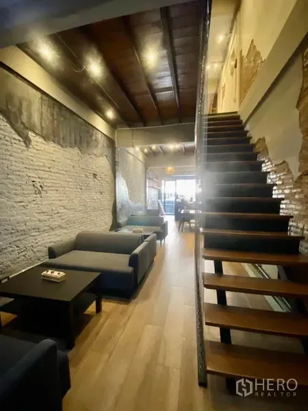 Shophouse for rent in Pathum Wan, Bangkok - Narrow lounge area with sofas along textured brick wall and open-tread wooden staircase to the mezzanine.