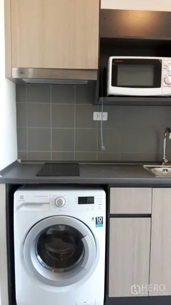 Condominium for rent in Samut Prakan - Compact kitchenette with induction cooktop, sink, microwave, gray tile backsplash, and a front-load washing machine.
