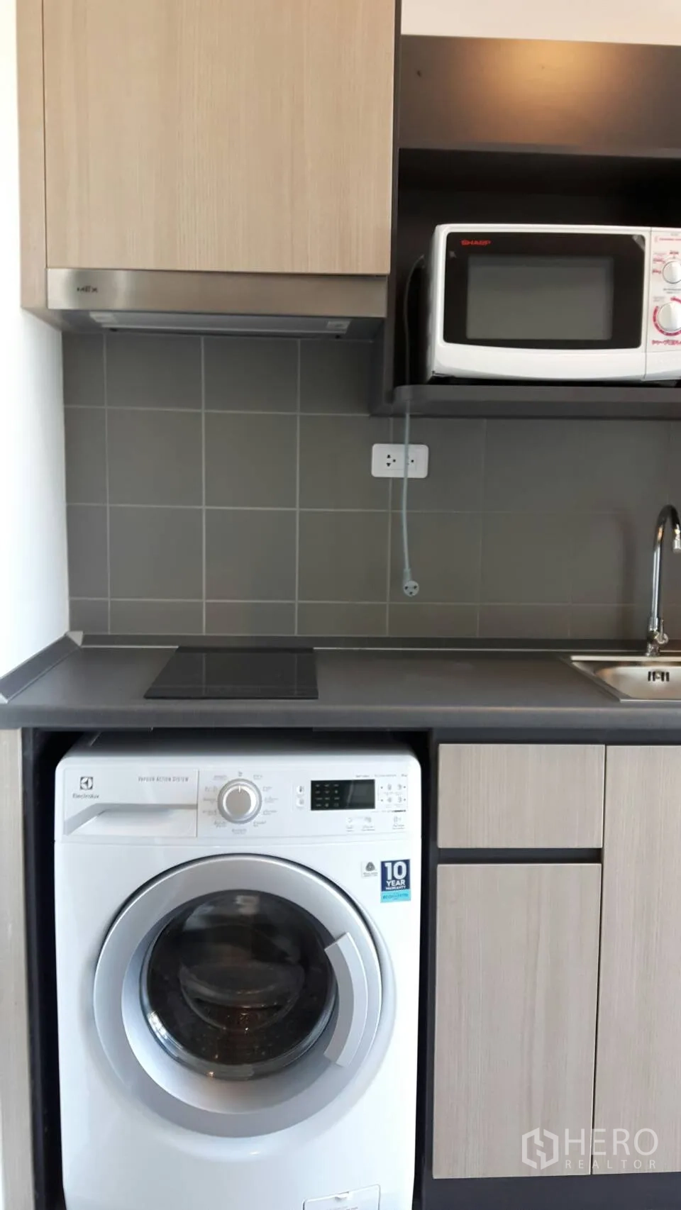 Condominium for rent in Samut Prakan - Compact kitchenette with induction cooktop, sink, microwave, gray tile backsplash, and a front-load washing machine.