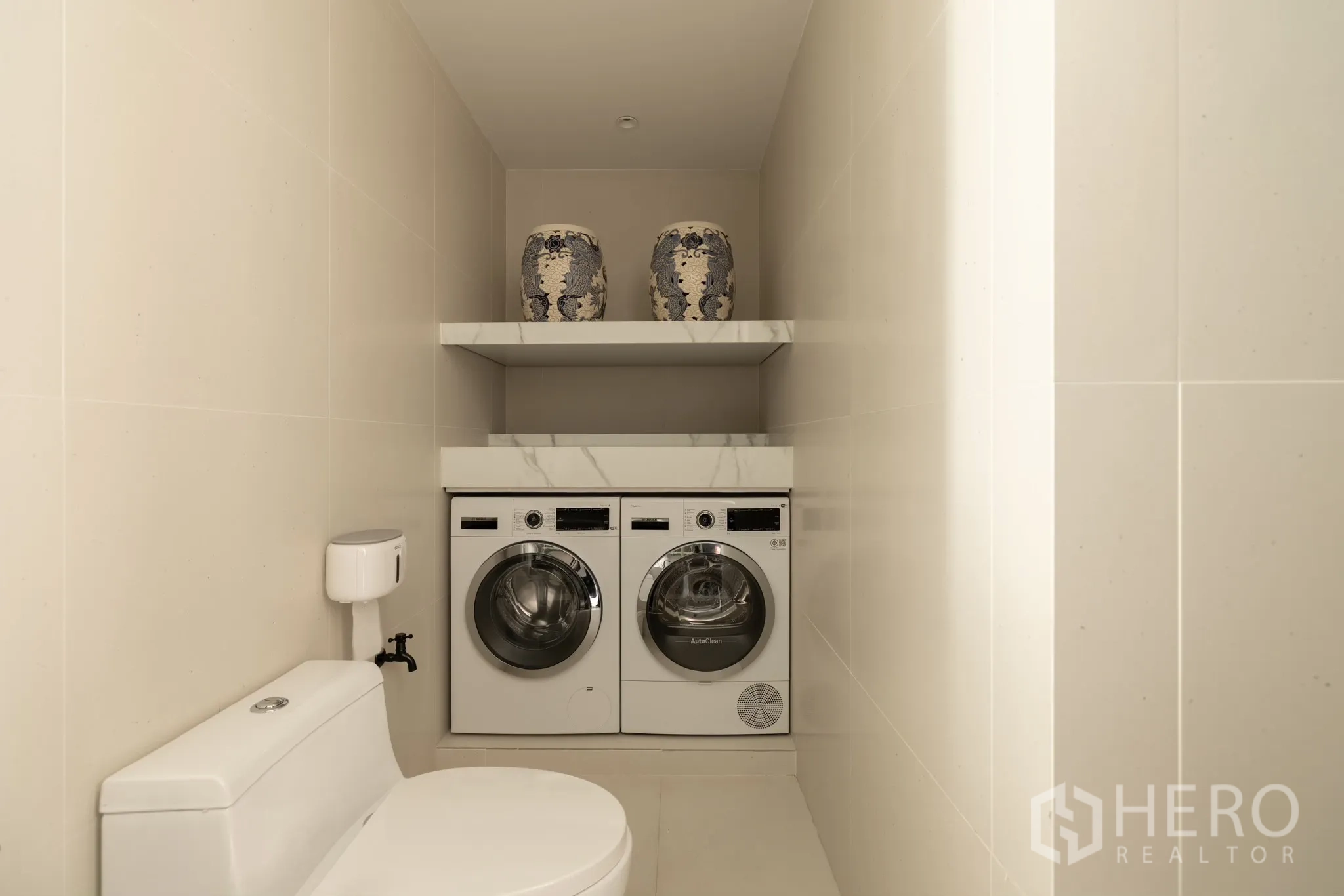 Condominium for sale in Khlong Toei, Bangkok - Compact utility room with washing machine, dryer and shelves beside a toilet.