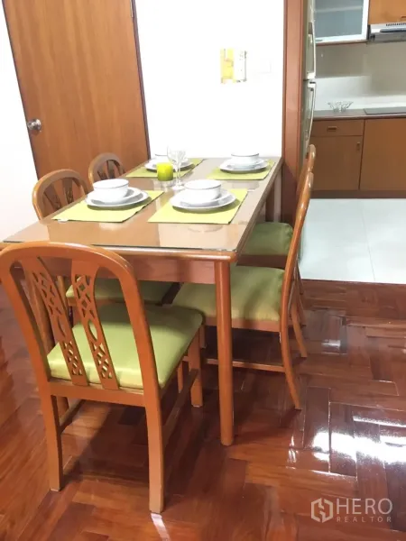 Condominium for rent in Watthana, Bangkok - Dining area with glass-top wooden table and green cushioned chairs beside the kitchen on polished parquet floors