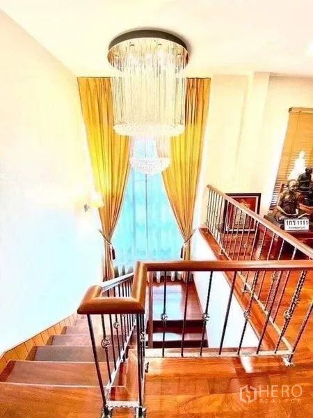 Detached House for sale in Saphan Sung, Bangkok - Wood staircase with metal balustrade beneath a crystal chandelier and tall window with drapes.