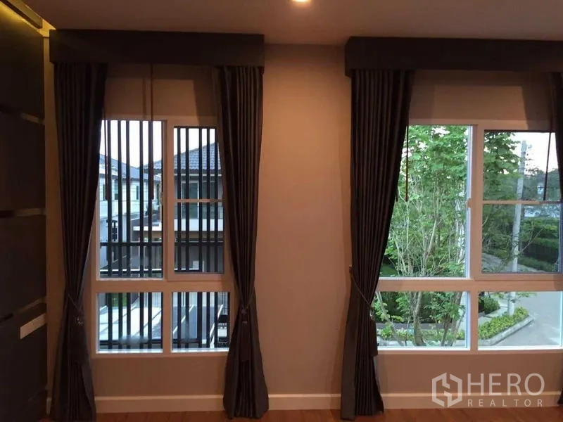 Townhouse for sale in Bang Khae, Bangkok - Two tall windows with dark curtains overlooking a landscaped community street.