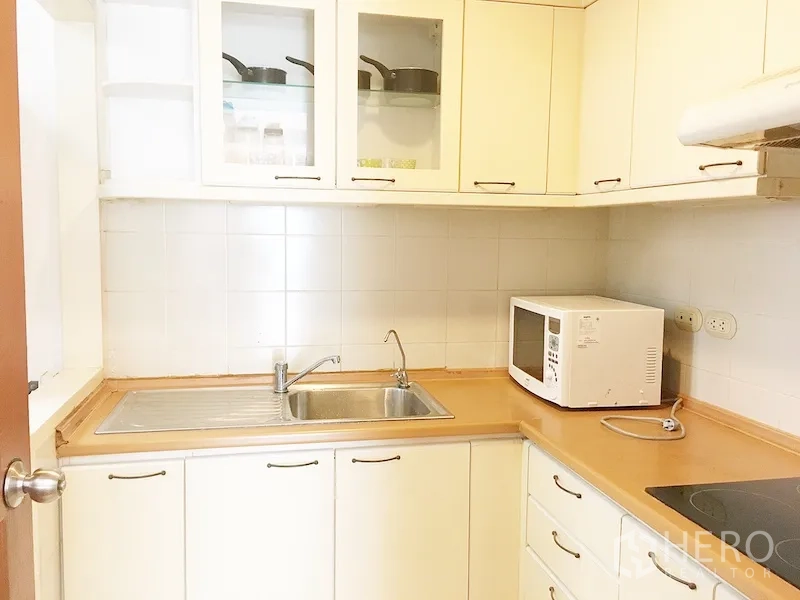 Condominium for rent in Pathum Wan, Bangkok - Compact white kitchen with L-shaped counter, sink, microwave, cooktop, and upper cabinets.