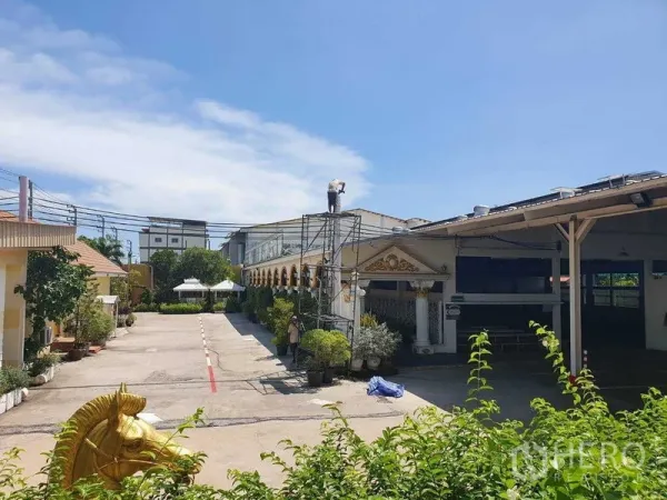 Warehouse for rent in Phra Pradaeng, Samut Prakan - Wide driveway between compound buildings with scaffolding and lush greenery.